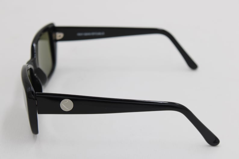 Puede incluir: Gafas de sol negras con montura rectangular y lentes oscuras. Las patillas son elegantes y presentan un emblema circular plateado. Las gafas de sol se encuentran sobre un fondo blanco liso, mostrando su dise&ntilde;o moderno.