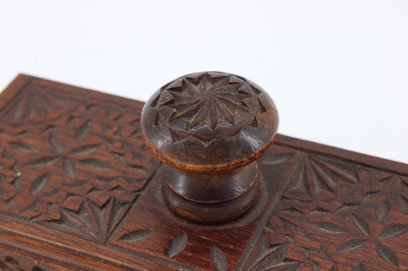May include: Close-up of a dark brown wooden inkwell with a carved floral design. The inkwell features a round, carved knob on top. The wood has a rich, warm tone, and the carving is detailed.