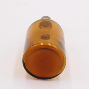 May include: Amber-colored glass bottle with a rounded body and a small, rounded stopper. The bottle has a smooth surface and a slightly darker base. The bottle is empty and appears to be vintage.