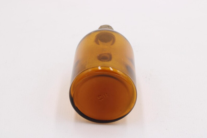 May include: Amber-colored glass bottle with a rounded body and a small, rounded stopper. The bottle has a smooth surface and a slightly darker base. The bottle is empty and appears to be vintage.