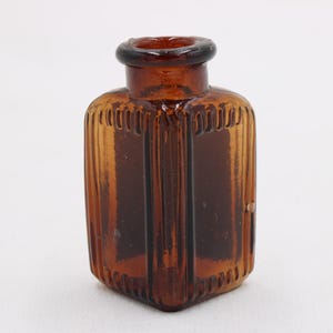 K&ouml;nnte beinhalten: Kleine, antike Flasche aus bernsteinfarbenem Glas mit quadratischem Boden und rundem Hals. Die Flasche hat vertikale Rippendetails an den Seiten und einen dunkelbraunen Rand. Die Flasche ist leer und steht vor einem wei&szlig;en Hintergrund.