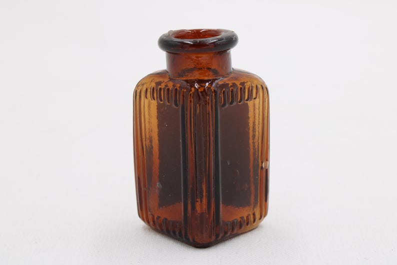 K&ouml;nnte beinhalten: Kleine, antike Flasche aus bernsteinfarbenem Glas mit quadratischem Boden und rundem Hals. Die Flasche hat vertikale Rippendetails an den Seiten und einen dunkelbraunen Rand. Die Flasche ist leer und steht vor einem wei&szlig;en Hintergrund.