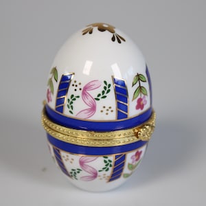 Può includere: Scatola portagioie a forma di uovo, bianca, con coperchio incernierato. Decorata con strisce blu, motivi floreali rosa e dettagli dorati. La scatola è alta circa 7.6 cm.