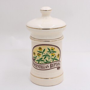 May include: Cream-colored ceramic apothecary jar with a lid and a gold rim. The jar features a decorative label with the text "Potentillas Reptans" and a botanical illustration of yellow flowers and green leaves.