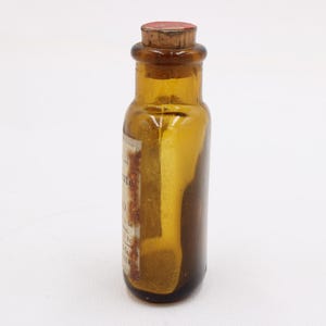 May include: Amber glass bottle with a cork stopper. The bottle has a label with faded text. The bottle is cylindrical with a rounded base and a narrow neck. The cork is reddish-brown.