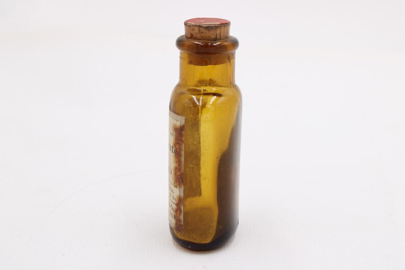 May include: Amber glass bottle with a cork stopper. The bottle has a label with faded text. The bottle is cylindrical with a rounded base and a narrow neck. The cork is reddish-brown.