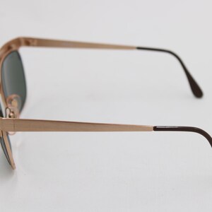 May include: Side view of gold-toned sunglasses with dark lenses. The frames are a light gold color, with dark brown earpieces. The sunglasses are set against a white background, highlighting their sleek design and classic style.