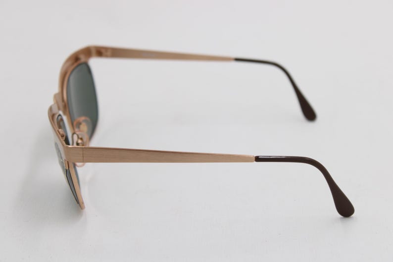 May include: Side view of gold-toned sunglasses with dark lenses. The frames are a light gold color, with dark brown earpieces. The sunglasses are set against a white background, highlighting their sleek design and classic style.