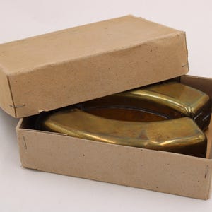 May include: Two brass ashtrays in a cardboard box. The ashtrays have a curved, rectangular shape with a shiny, golden finish. The box is open, revealing the ashtrays inside. The box is made of plain, light brown cardboard.