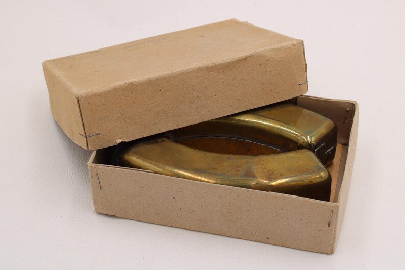 May include: Two brass ashtrays in a cardboard box. The ashtrays have a curved, rectangular shape with a shiny, golden finish. The box is open, revealing the ashtrays inside. The box is made of plain, light brown cardboard.