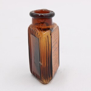 K&ouml;nnte beinhalten: Eine Vintage-Flasche aus bernsteinfarbenem Glas mit quadratischer Form und abgerundeter Oberseite. Die Flasche hat vertikale Rippendetails an den Seiten und scheint Text auf dem Etikett zu haben. Die Flasche ist leer und steht auf einer wei&szlig;en Oberfl&auml;che.
