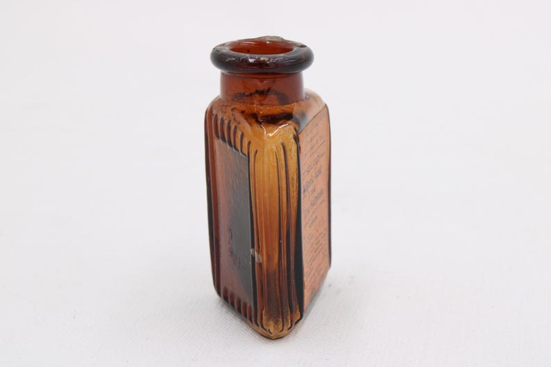 K&ouml;nnte beinhalten: Eine Vintage-Flasche aus bernsteinfarbenem Glas mit quadratischer Form und abgerundeter Oberseite. Die Flasche hat vertikale Rippendetails an den Seiten und scheint Text auf dem Etikett zu haben. Die Flasche ist leer und steht auf einer wei&szlig;en Oberfl&auml;che.