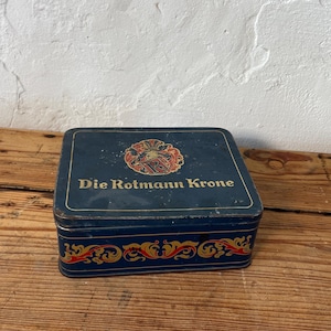 May include: A vintage, rectangular tin box with a dark blue background. The box features the text "Die Rotmann Krone" in gold lettering, along with a decorative emblem. The edges are adorned with red and gold scrollwork.
