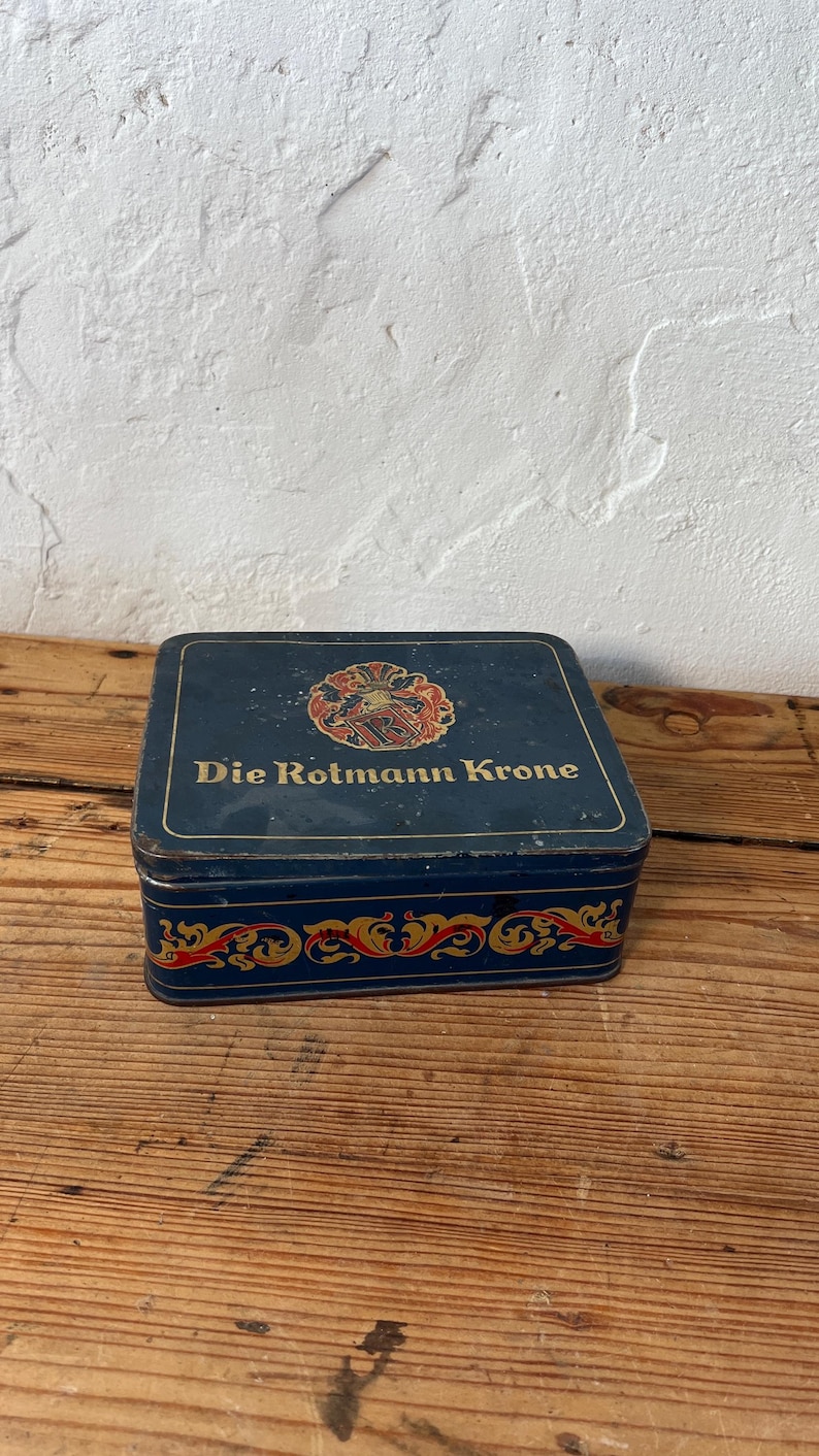 May include: A vintage, rectangular tin box with a dark blue background. The box features the text "Die Rotmann Krone" in gold lettering, along with a decorative emblem. The edges are adorned with red and gold scrollwork.