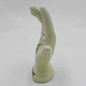 May include: A pale green ceramic hand sculpture with black line drawings and text. The hand is in a curved position, with the fingers pointing upwards. The base of the hand has text on it.