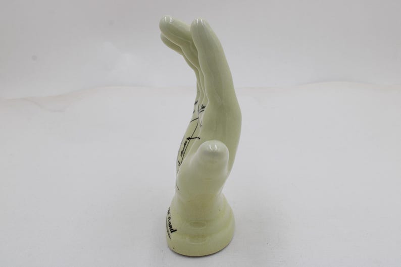 May include: A pale green ceramic hand sculpture with black line drawings and text. The hand is in a curved position, with the fingers pointing upwards. The base of the hand has text on it.