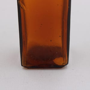 May include: Close-up of a brown glass bottle with a rectangular shape. The bottle has a dark amber color and appears to be empty. The bottom of the bottle has a slightly rounded edge.