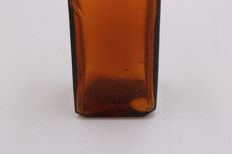 May include: Close-up of a brown glass bottle with a rectangular shape. The bottle has a dark amber color and appears to be empty. The bottom of the bottle has a slightly rounded edge.