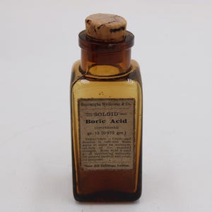 May include: An antique amber glass bottle with a cork stopper. The label reads "SOLOID Boric Acid" and includes directions and measurements of 0.972 grams. The bottle is rectangular with rounded corners.