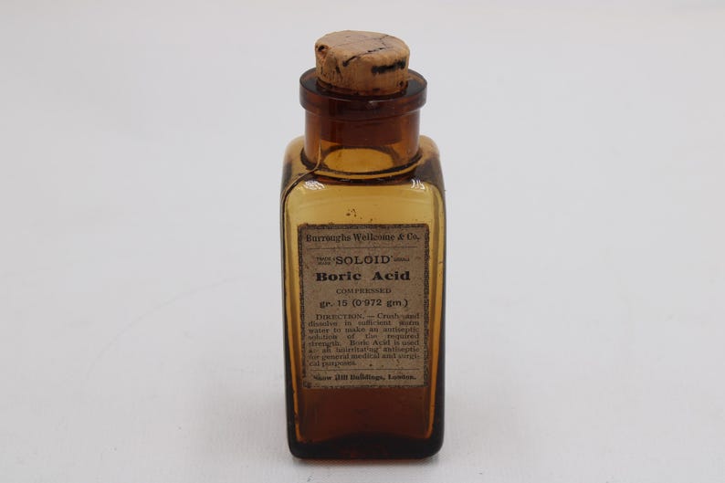 May include: An antique amber glass bottle with a cork stopper. The label reads "SOLOID Boric Acid" and includes directions and measurements of 0.972 grams. The bottle is rectangular with rounded corners.