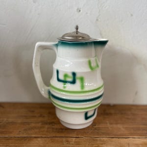 May include: White ceramic pitcher with a silver lid. The pitcher features green and blue geometric designs and stripes. The handle is white, and the lid has a small decorative top. The pitcher is on a wooden surface.