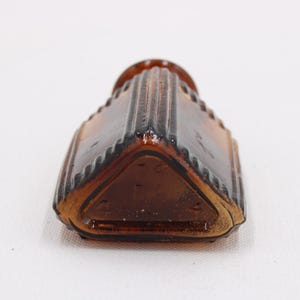 K&ouml;nnte beinhalten: Eine Vintage-Flasche aus braunem Glas mit einer einzigartigen, geometrischen Form. Die Flasche hat ein geriffeltes Design an den Seiten und eine dreieckige Basis. Die Farbe der Flasche ist ein tiefes Braun, und sie scheint leer zu sein.