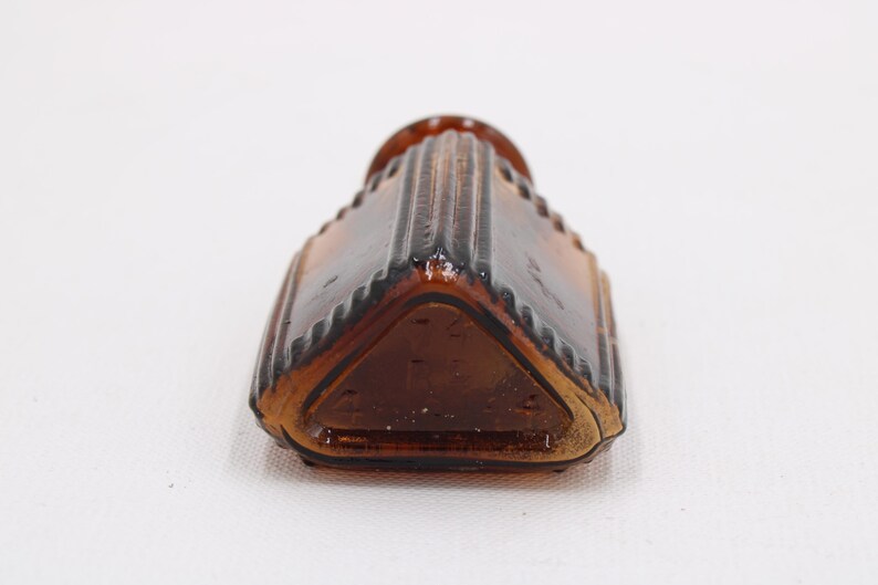 K&ouml;nnte beinhalten: Eine Vintage-Flasche aus braunem Glas mit einer einzigartigen, geometrischen Form. Die Flasche hat ein geriffeltes Design an den Seiten und eine dreieckige Basis. Die Farbe der Flasche ist ein tiefes Braun, und sie scheint leer zu sein.