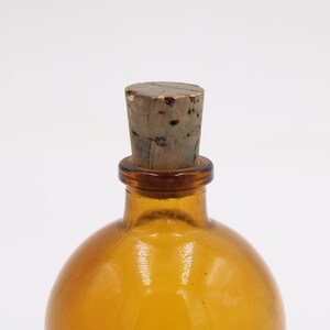 May include: Amber-colored glass bottle with a cork stopper. The bottle has a rounded shape and a narrow neck. The cork is a light brown color and appears to be made of wood. The bottle is empty and sits against a white background.