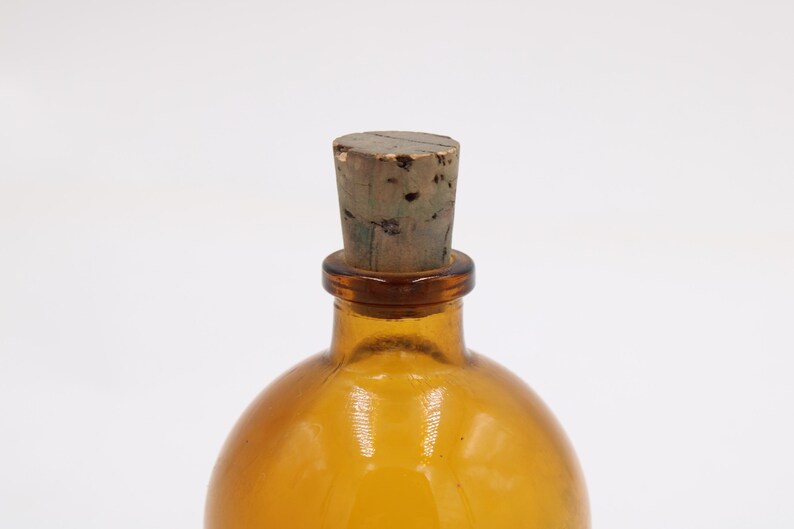 May include: Amber-colored glass bottle with a cork stopper. The bottle has a rounded shape and a narrow neck. The cork is a light brown color and appears to be made of wood. The bottle is empty and sits against a white background.