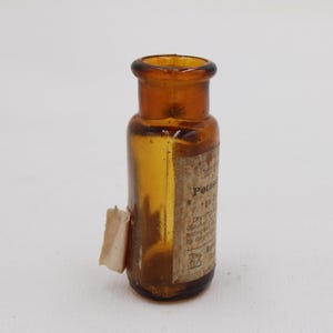 Puede incluir: Peque&ntilde;a botella de vidrio color &aacute;mbar con etiqueta. La botella tiene base redondeada y cuello estrecho. La etiqueta parece tener texto. Probablemente un art&iacute;culo antiguo o vintage.