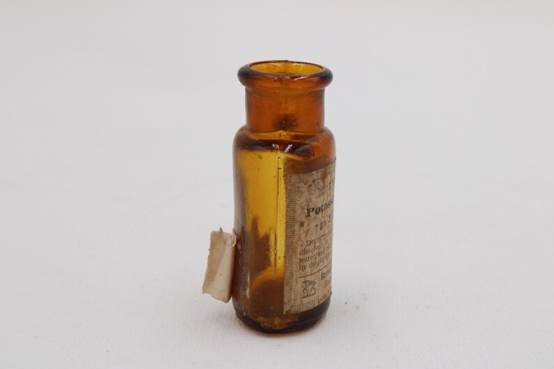 Puede incluir: Peque&ntilde;a botella de vidrio color &aacute;mbar con etiqueta. La botella tiene base redondeada y cuello estrecho. La etiqueta parece tener texto. Probablemente un art&iacute;culo antiguo o vintage.