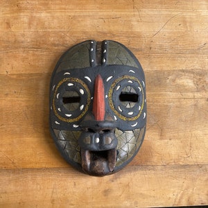 May include: A wooden mask with a black, red, white and gold geometric design. The mask has two circular eye holes and a large, open mouth.