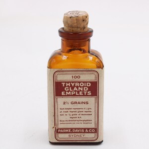 May include: An amber glass bottle with a cork stopper, labeled "100 Thyroid Gland Emplets, 2 1/2 Grains." The label is cream-colored with maroon text. The bottle is likely an antique pharmaceutical container.