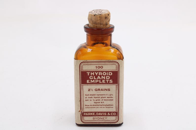 May include: An amber glass bottle with a cork stopper, labeled "100 Thyroid Gland Emplets, 2 1/2 Grains." The label is cream-colored with maroon text. The bottle is likely an antique pharmaceutical container.