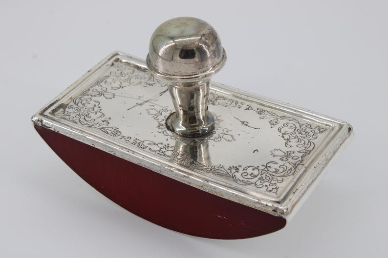 May include: A vintage silver ink blotter with a red base. The rectangular blotter has a decorative floral pattern on the silver top and a rounded, curved base. The handle is a rounded silver knob.