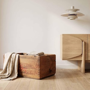 Può includere: Una cassa rettangolare in legno con venature e giunzioni a pettine visibili, drappeggiata con un tessuto beige. Accanto si trova un grande vaso in ceramica color bianco sporco con corpo largo e collo stretto, vicino a una credenza in rovere.