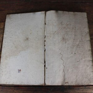 以下が含まれることがあります： 開かれたアンティーク本。黄ばんだページに手書きの文字と波線が描かれています。本は、ヴィンテージ感を際立たせる、素朴な濃い木製の表面に置かれています。