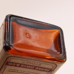 May include: Close-up of a vintage amber glass bottle. The bottom of the bottle has the number "907" embossed. The label reads "PARKE, DAVIS & CO. SYDNEY". The bottle has a rectangular shape with rounded corners.