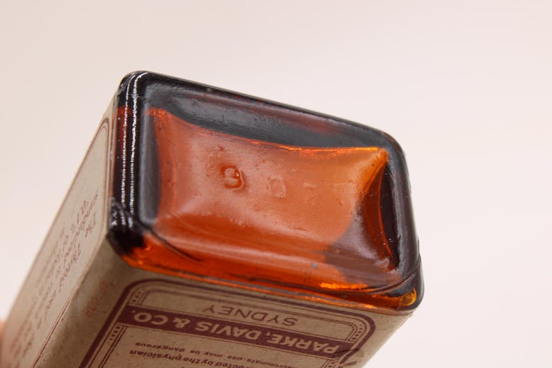 May include: Close-up of a vintage amber glass bottle. The bottom of the bottle has the number "907" embossed. The label reads "PARKE, DAVIS & CO. SYDNEY". The bottle has a rectangular shape with rounded corners.
