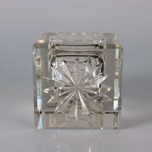 May include: Clear, square crystal inkwell with a starburst design on the front. The inkwell has a small, round opening on top and is made of faceted glass. The design is symmetrical and reflects light.