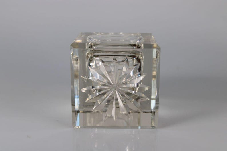 May include: Clear, square crystal inkwell with a starburst design on the front. The inkwell has a small, round opening on top and is made of faceted glass. The design is symmetrical and reflects light.