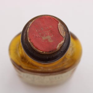 May include: An overhead shot of a vintage bottle, featuring a red wax seal. The bottle has a rounded form and a brown neck. The seal is partially damaged, showing the bottle's age. The bottle's body is a light amber colour.