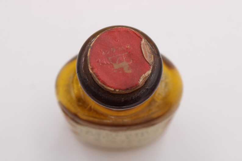 May include: An overhead shot of a vintage bottle, featuring a red wax seal. The bottle has a rounded form and a brown neck. The seal is partially damaged, showing the bottle's age. The bottle's body is a light amber colour.