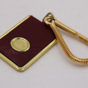 May include: A rectangular burgundy leather keychain with a gold border and a gold oval emblem. The keychain has a gold spring-like loop for attaching keys. The emblem features a stylized design.