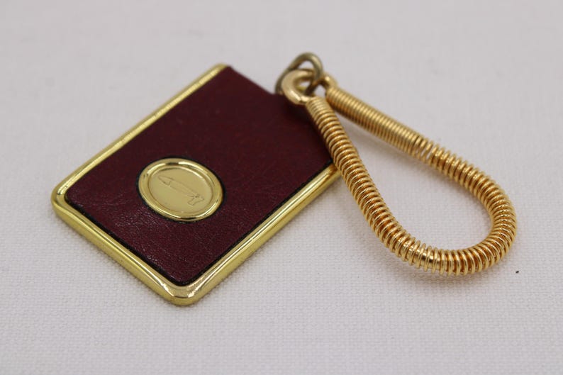 May include: A rectangular burgundy leather keychain with a gold border and a gold oval emblem. The keychain has a gold spring-like loop for attaching keys. The emblem features a stylized design.