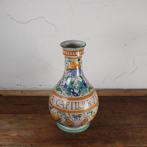 May include: A decorative ceramic vase with a bulbous body and a narrow neck. The vase features a colorful floral and leaf design in blue, yellow, and green, with the text "CAPILOR RII" in a band around the middle. The base and rim are green.