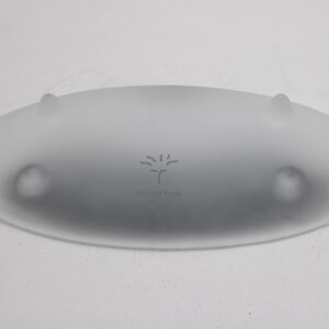 K&ouml;nnte beinhalten: Ovale Servierschale aus mattiertem Glas mit drei kleinen, abgerundeten F&uuml;&szlig;en. Die Schale hat einen leicht erh&ouml;hten Rand und eine subtile, strukturierte Oberfl&auml;che. Der Text "MARGOT WOHL" und ein stilisiertes Baumdesign sind in die Mitte ge&auml;tzt.