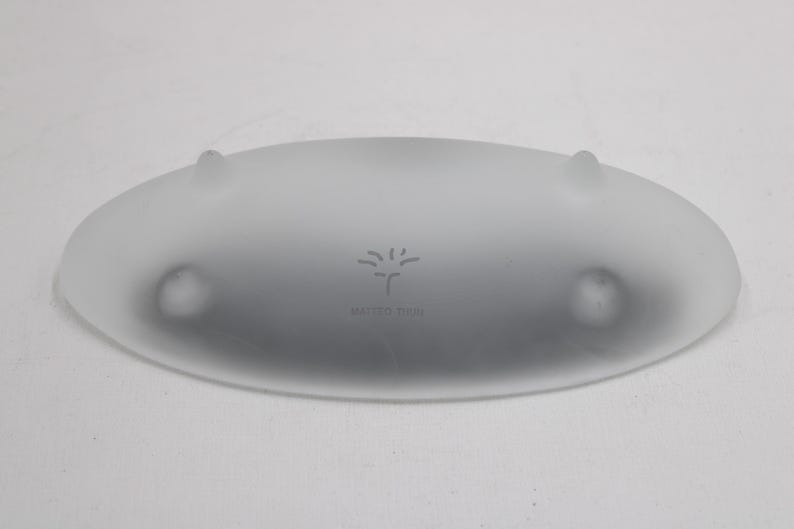K&ouml;nnte beinhalten: Ovale Servierschale aus mattiertem Glas mit drei kleinen, abgerundeten F&uuml;&szlig;en. Die Schale hat einen leicht erh&ouml;hten Rand und eine subtile, strukturierte Oberfl&auml;che. Der Text "MARGOT WOHL" und ein stilisiertes Baumdesign sind in die Mitte ge&auml;tzt.