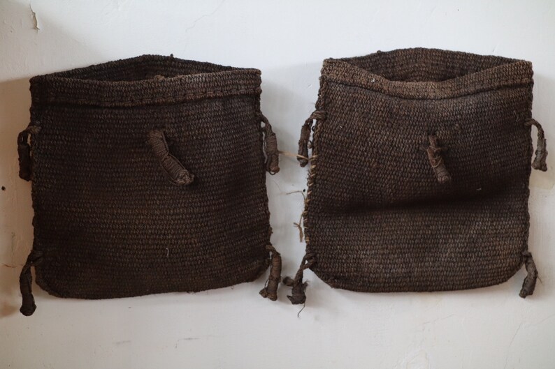 May include: Two brown woven bags with drawstrings hanging on a white wall.