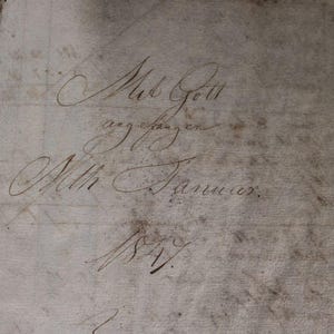 以下が含まれることがあります： 茶色のインクで書かれた、色あせた手書き文字が書かれた開いた本のページ。テキストには「Med Gott」と日付「1857」が含まれています。紙は使い古された、古くなった外観をしており、アンティークの文書であることを示唆しています。
